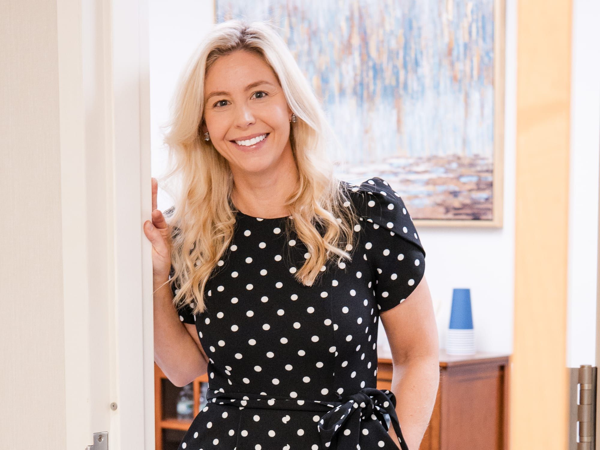 A woman with long blonde hair in a black and white polka-dot dress, smiling and leaning in a wooden doorway, with an abstract painting behind her
