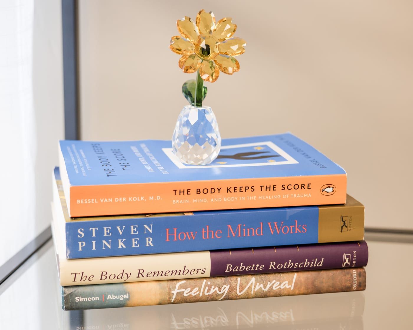 A stack of four books on a glass shelf with a small glass vase and a yellow flower on top