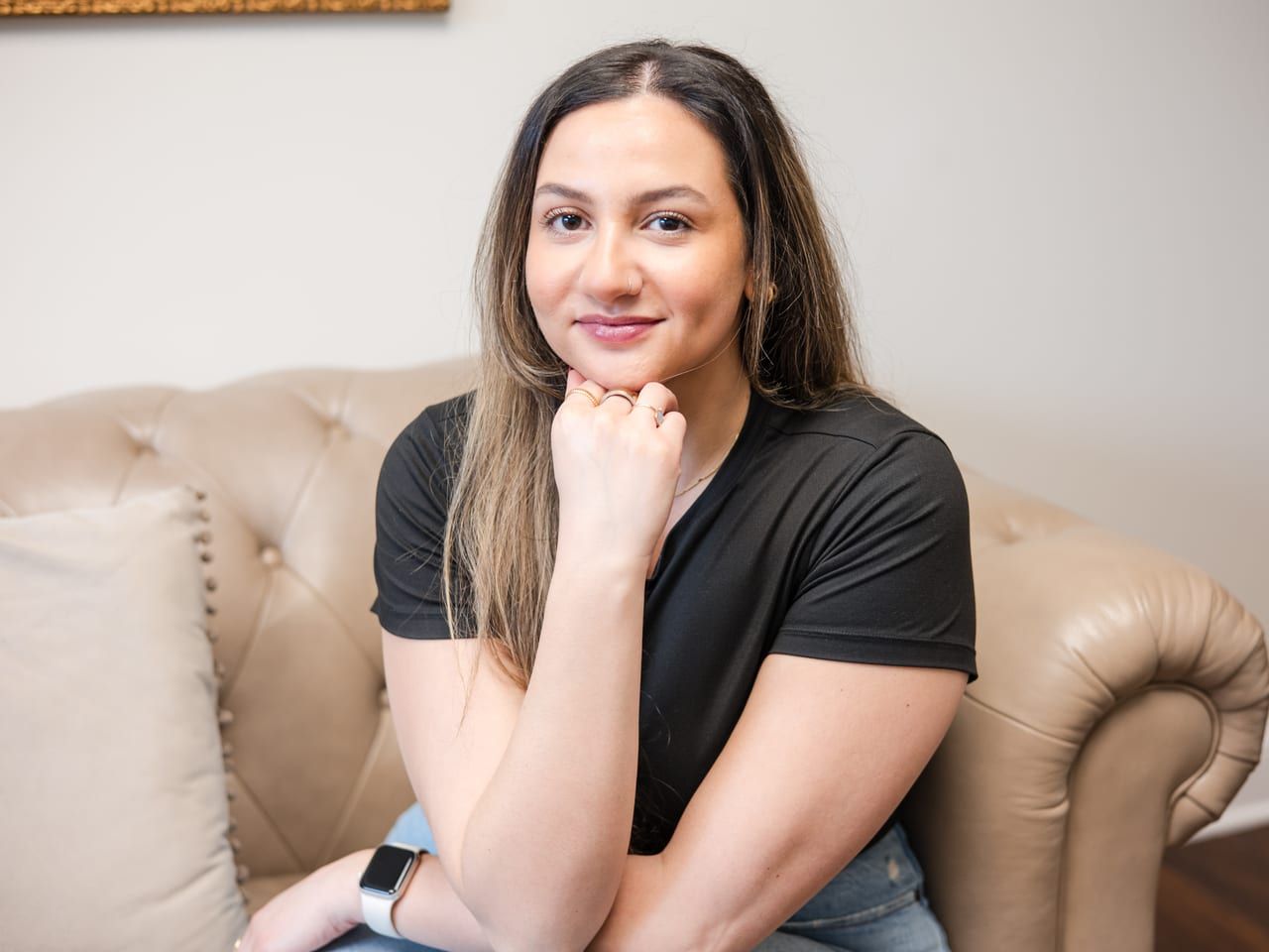 Carly Frascino seated on a beige leather sofa in a black tee, smiling with her hand at her chin
