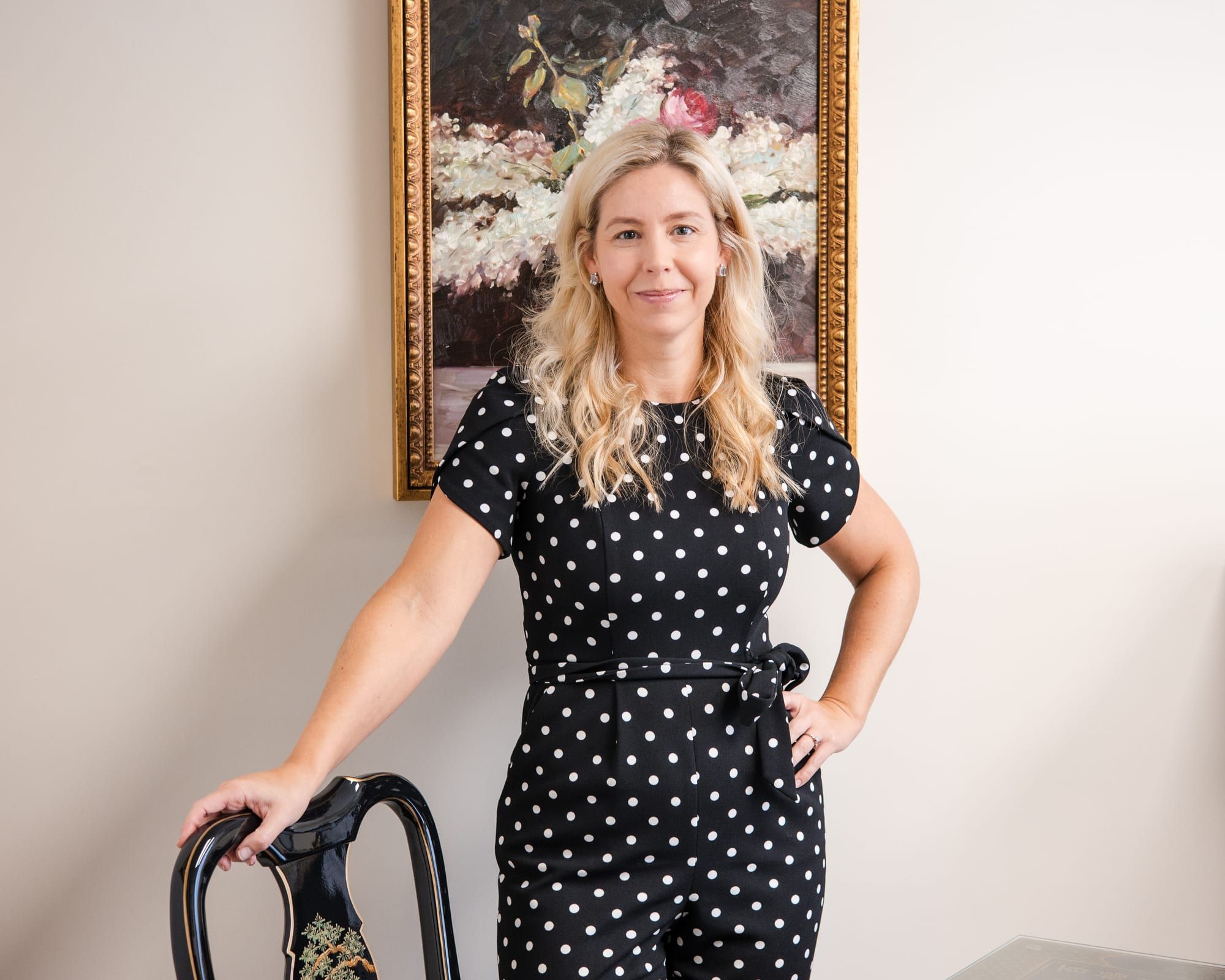 A woman with long blonde hair in a black and white polka-dot jumpsuit, standing with a hand on her hip behind a black chair, a framed floral painting behind her