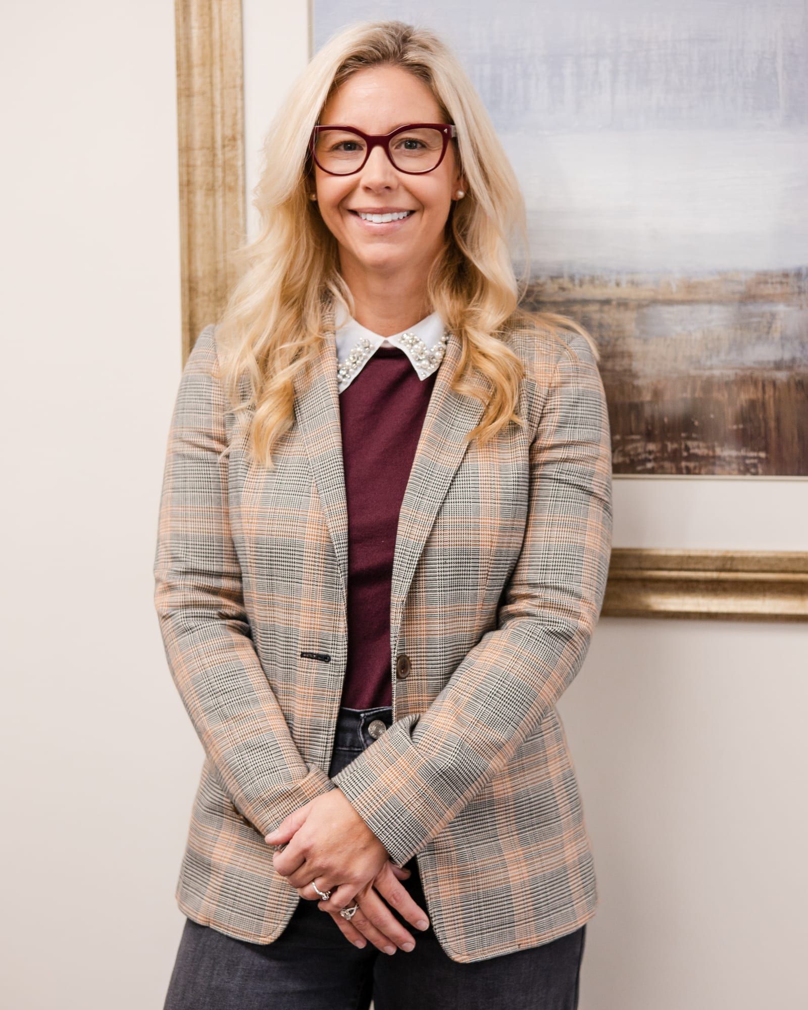 A woman with long blonde hair and red-framed glasses in a plaid blazer over a burgundy top, smiling beside a framed landscape painting