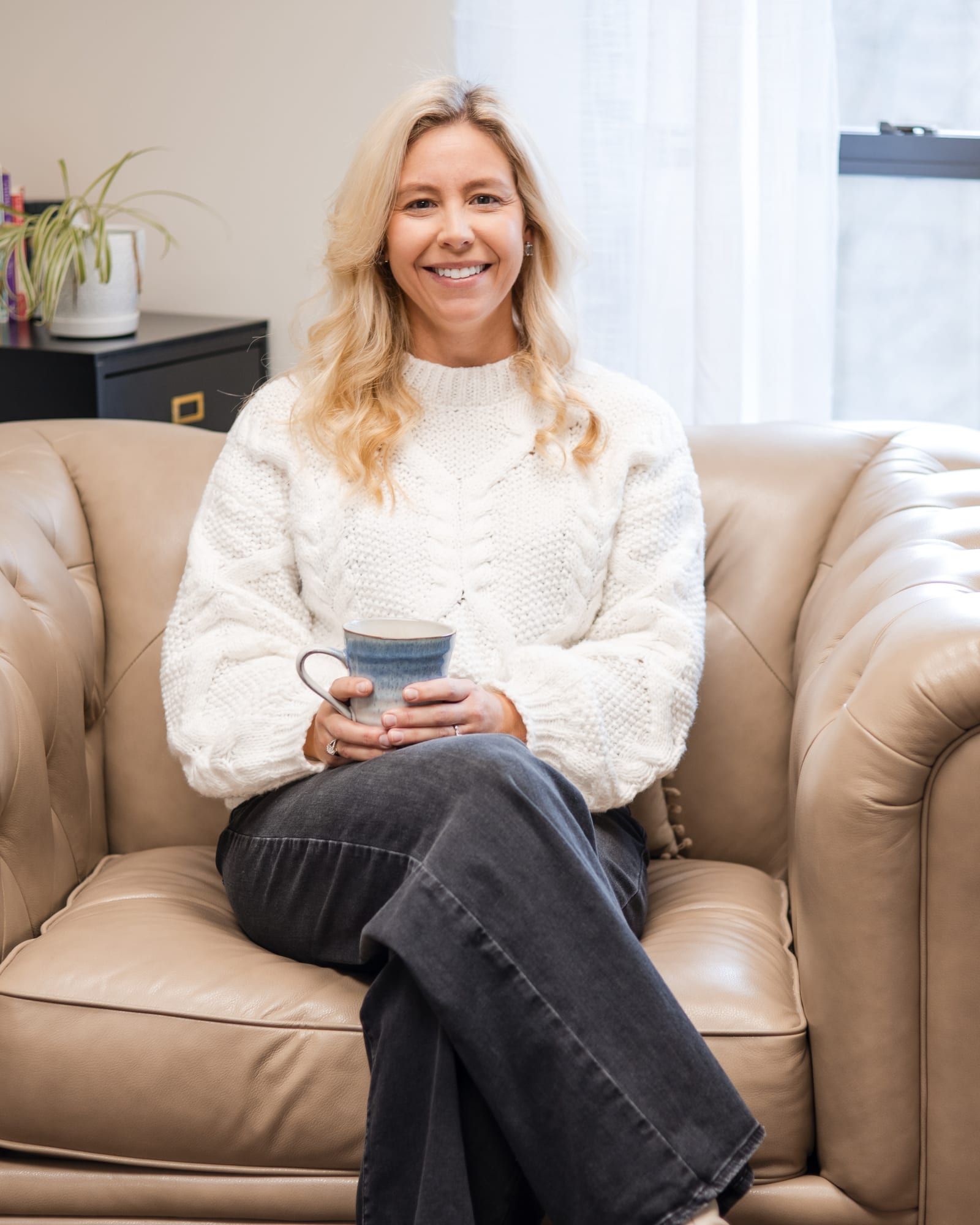 A woman with long blonde hair in a white knit sweater, smiling and holding a mug in a beige leather armchair beside a window