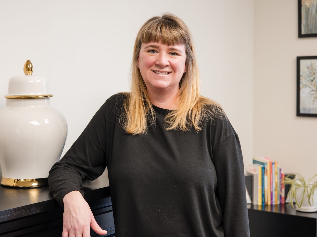 Lindsay Thompson Neill in a black long-sleeve top, smiling and leaning on a black console beside a white ginger jar