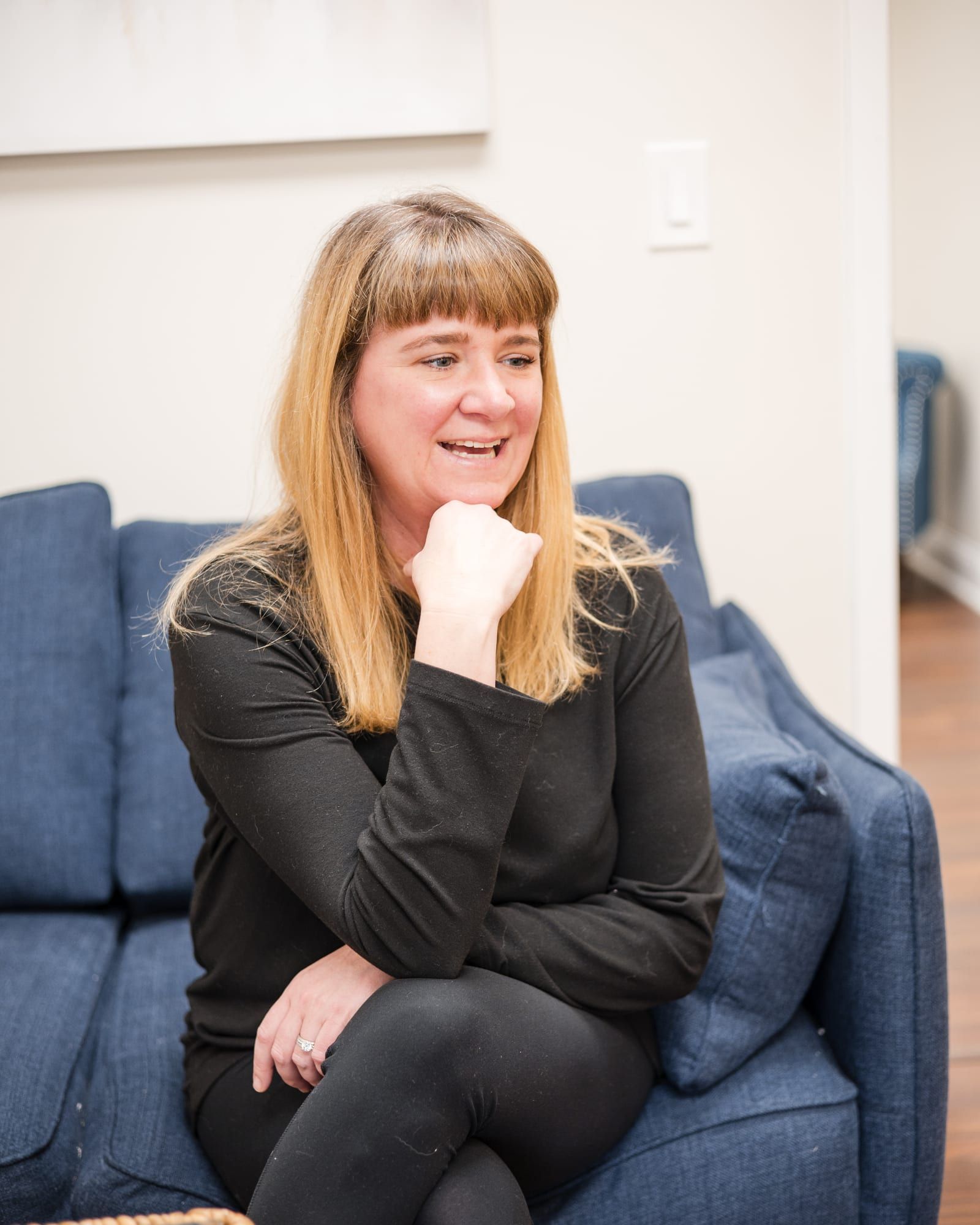 Lindsay Thompson Neill seated on a blue sofa in a black long-sleeve top, smiling with her chin on her hand