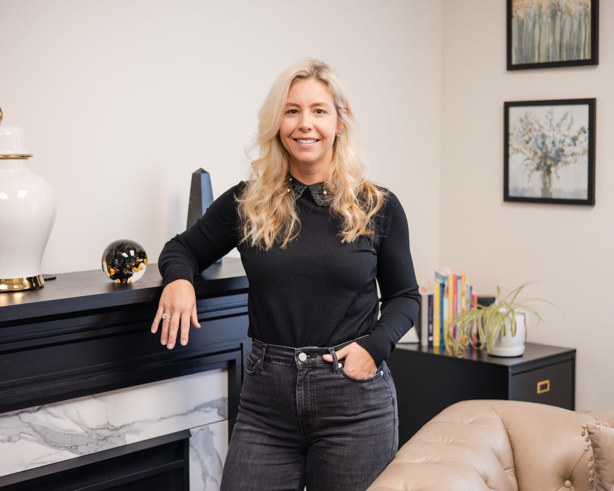A woman with long blonde hair in a black sweater, leaning on a black fireplace console with a marble front, framed art and a bookshelf behind her