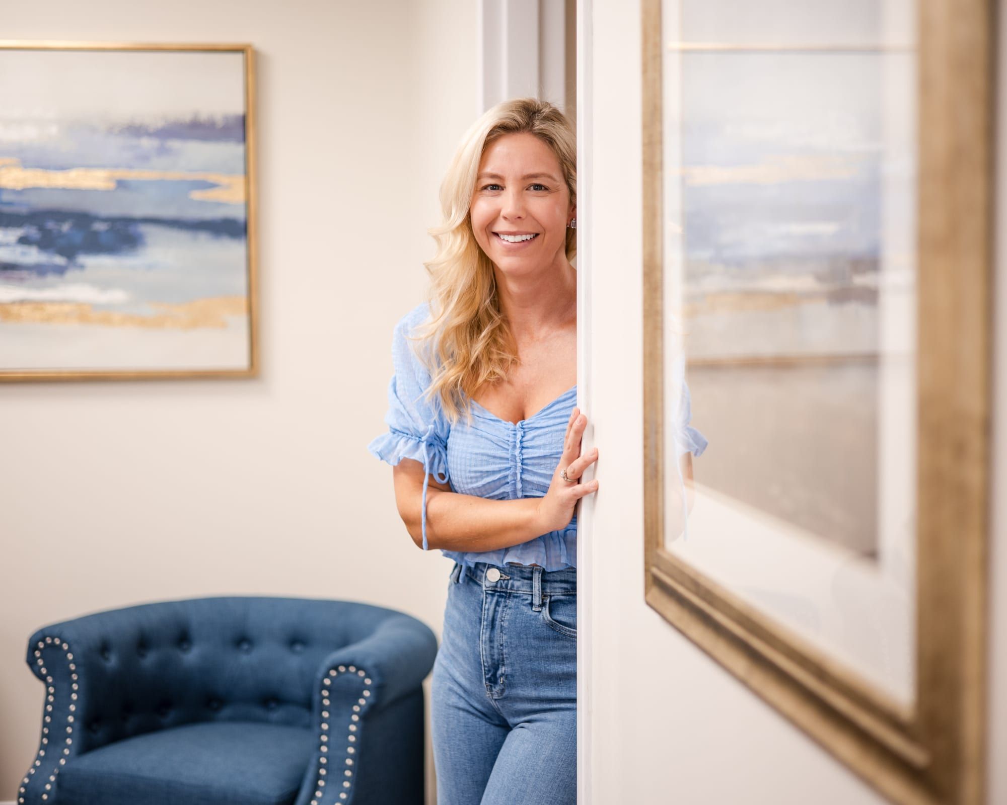 A woman with long blonde hair in a light blue top and jeans, smiling and leaning on a door frame beside a blue tufted armchair and a framed painting