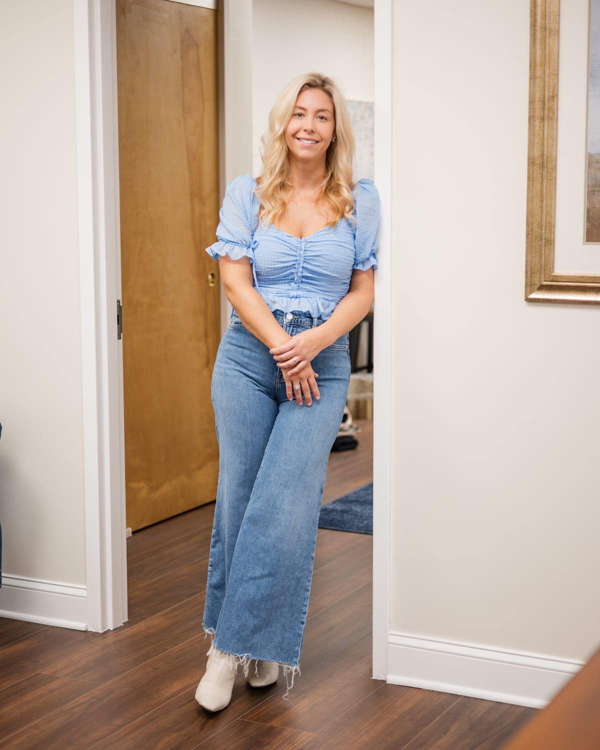 A woman with long blonde hair smiling in a wooden doorway with her hands clasped, in a light blue ruched top and wide-leg jeans