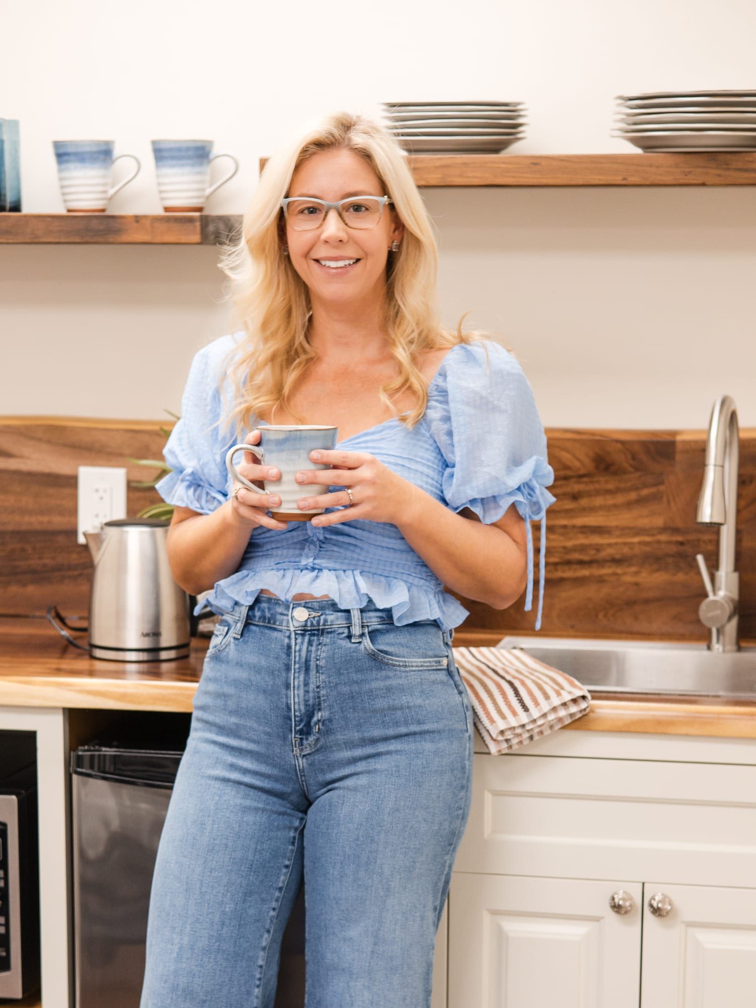 A woman with long blonde hair and glasses in a light blue top and wide-leg jeans, smiling and holding a mug in the office kitchen