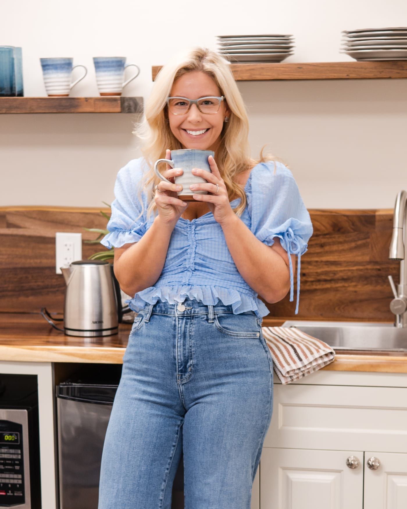 Lindsey Capelli smiling and holding a blue and white mug in the office kitchen, in a light blue top and glasses