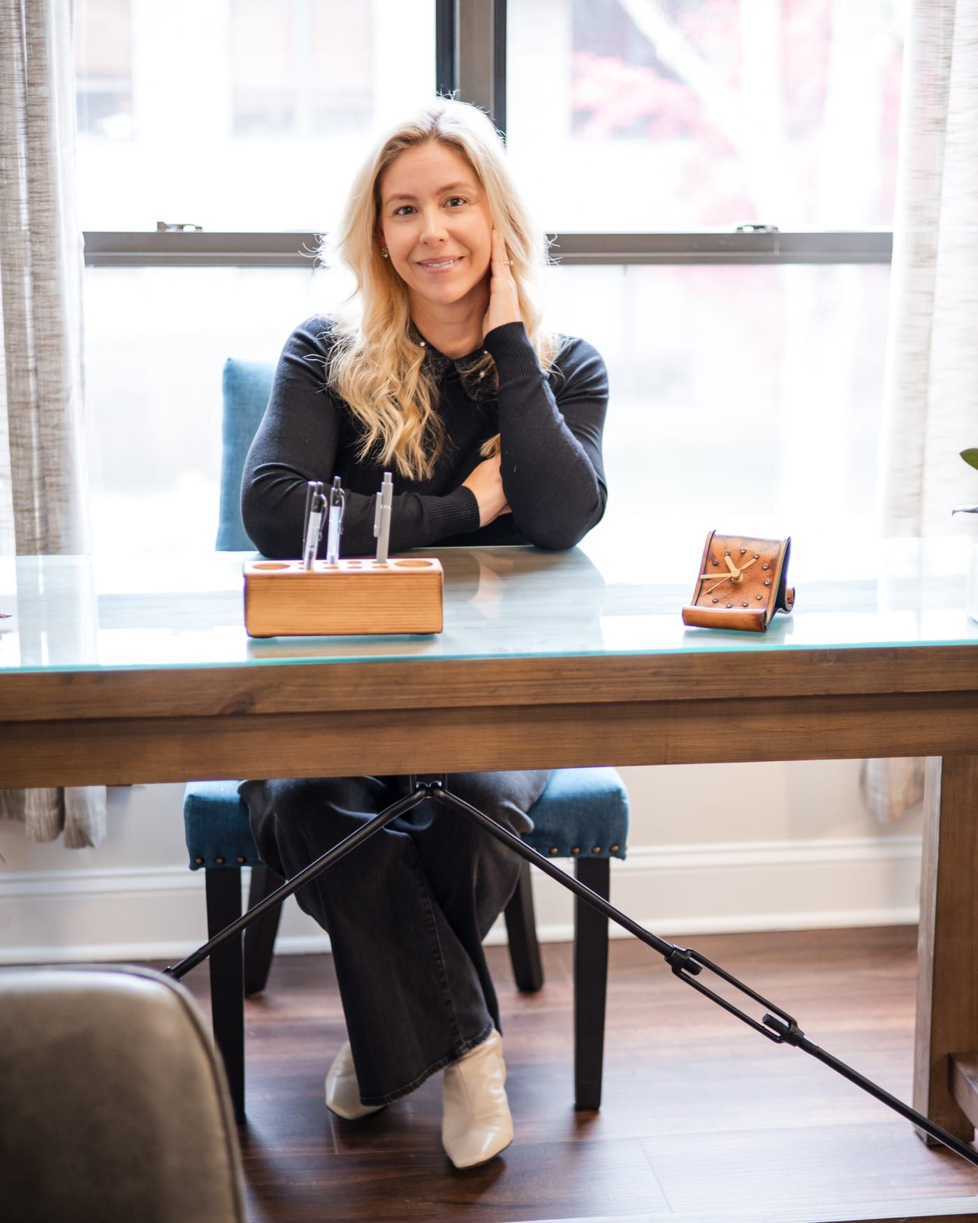 A woman with long blonde hair in a black sweater, seated at a wooden desk by a bright window, one hand raised to her neck