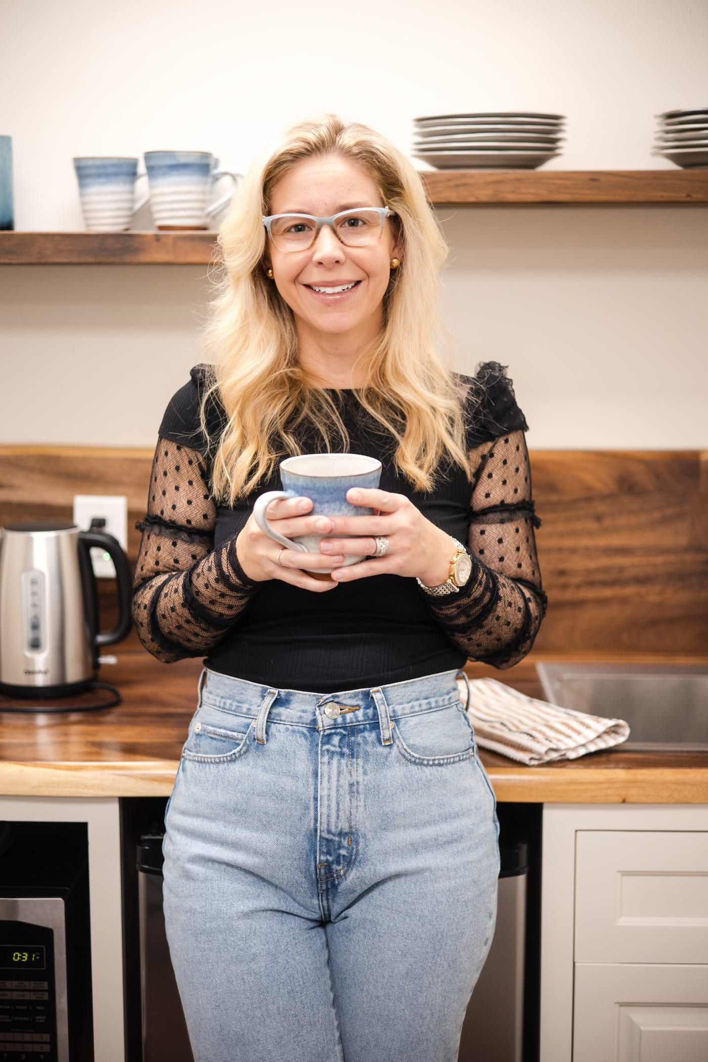 A woman with long blonde hair and glasses smiling in the office kitchen, holding a blue and white mug in both hands, in a black top with dotted sheer sleeves and light jeans