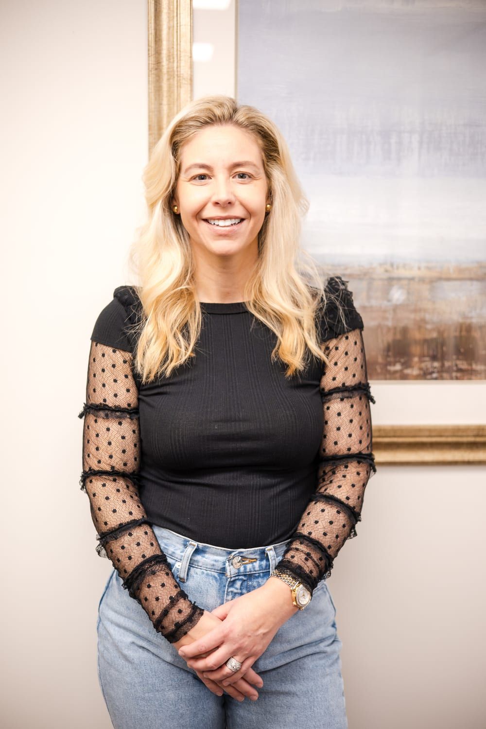 A woman with long blonde hair smiling with her hands clasped in front of her, in a black top with dotted sheer sleeves and light jeans, in front of a framed painting