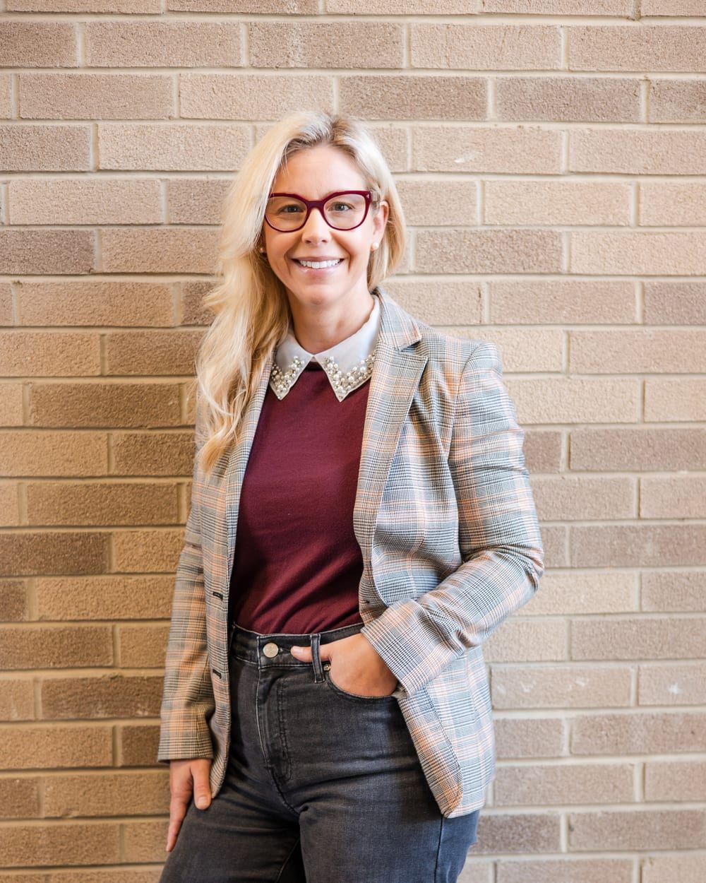A woman with long blonde hair and red-framed glasses smiling with one hand in her pocket, in a plaid blazer and a burgundy top, in front of a tan brick wall