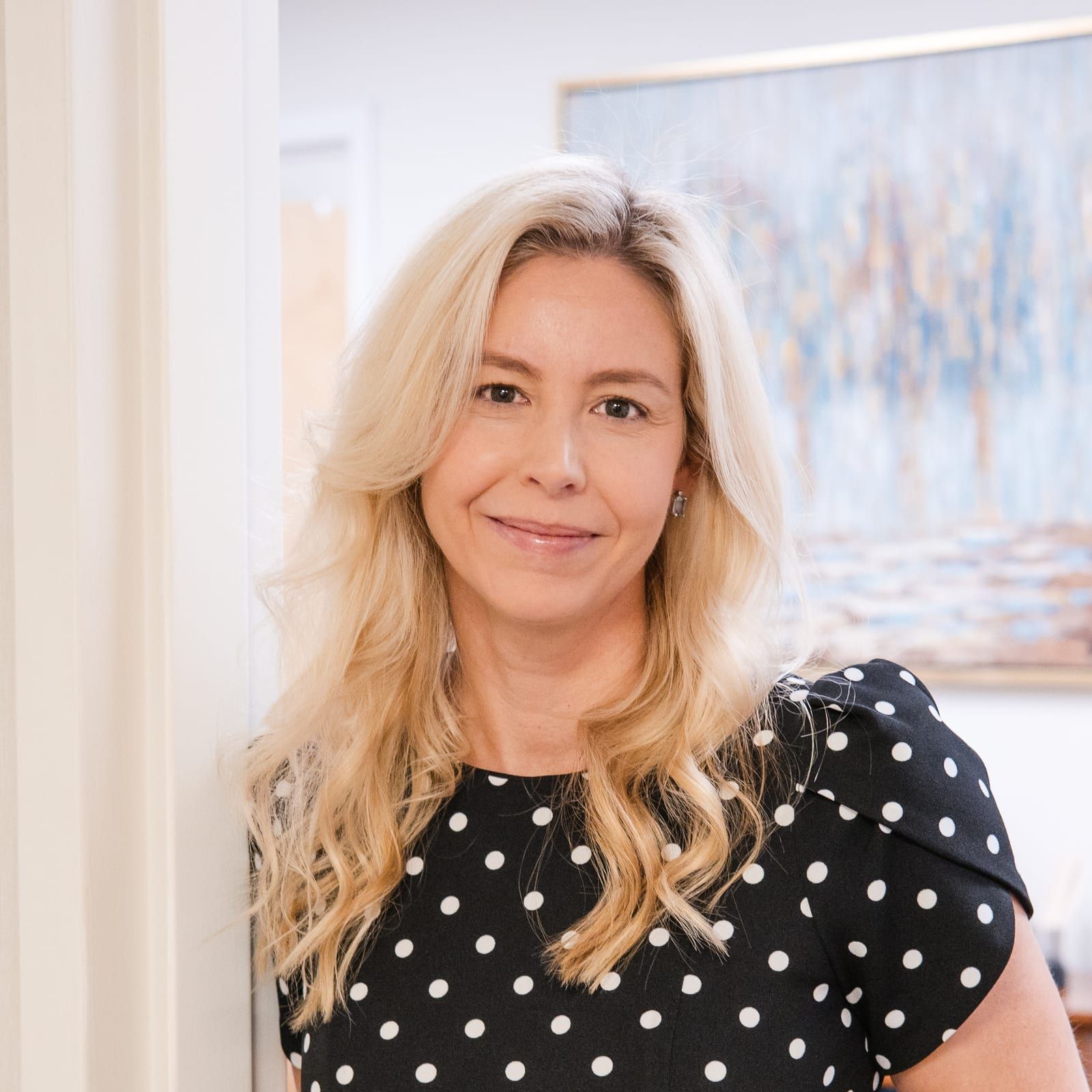 A woman with long blonde hair smiling in a black and white polka-dot jumpsuit, leaning in an office doorway