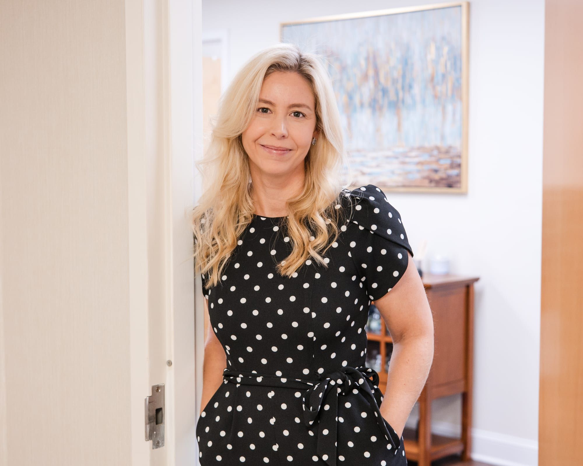 A woman with long blonde hair in a black and white polka-dot jumpsuit, smiling and leaning in a wooden doorway