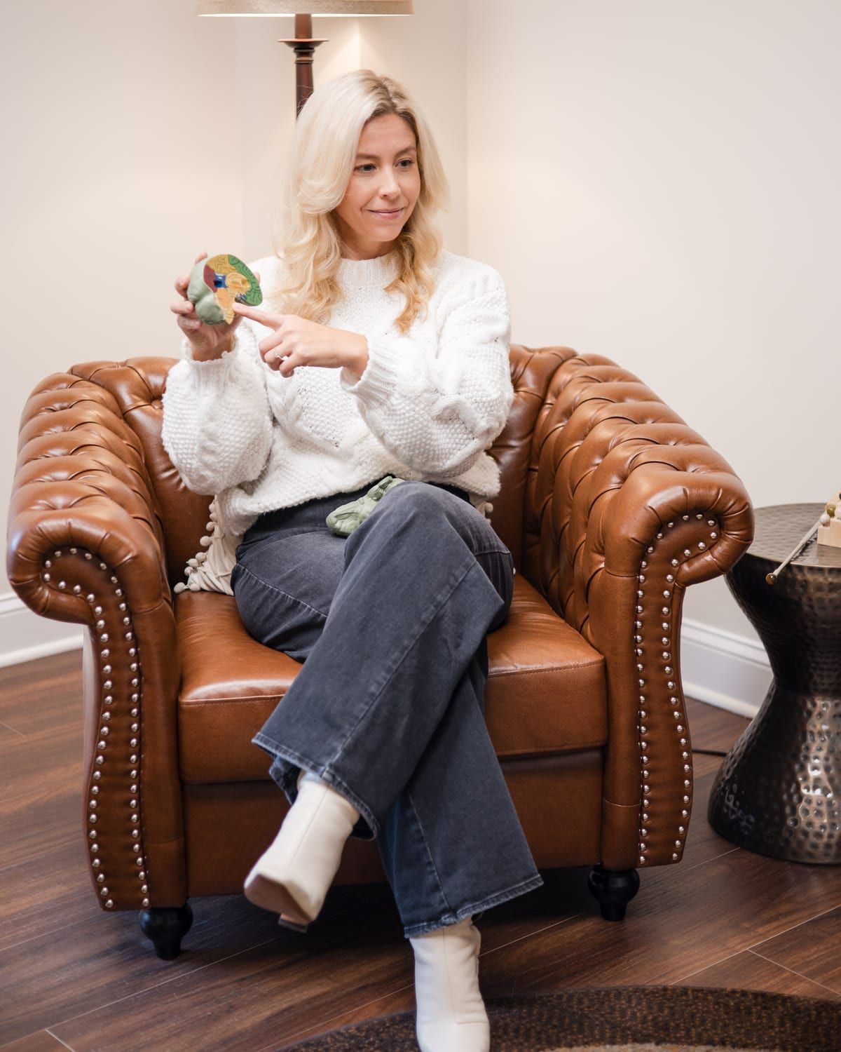 A woman in a white knit sweater smiling in a brown leather armchair, holding a color-coded brain model in one hand and pointing to it with the other