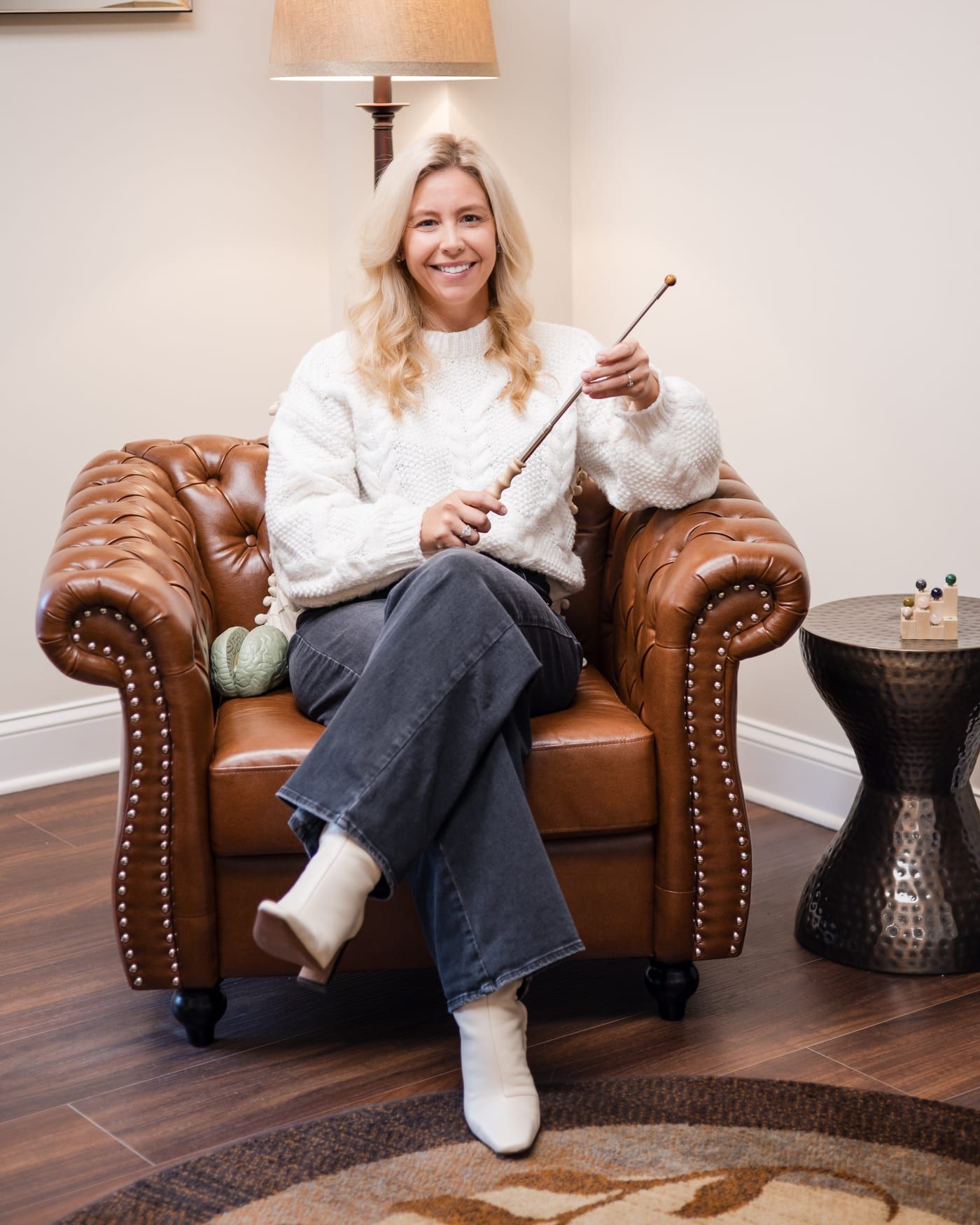 Lindsey Capelli smiling in a brown leather tufted armchair, in a white knit sweater, holding a slim wooden pointer across her lap