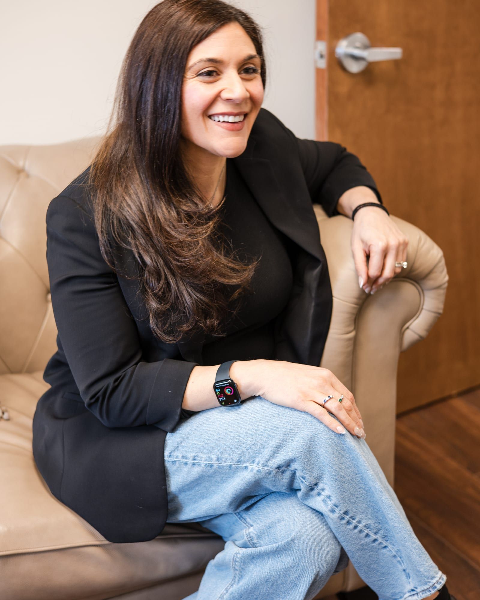 Marina Fried seated in a beige leather chair in a black blazer and jeans, smiling and looking to the side