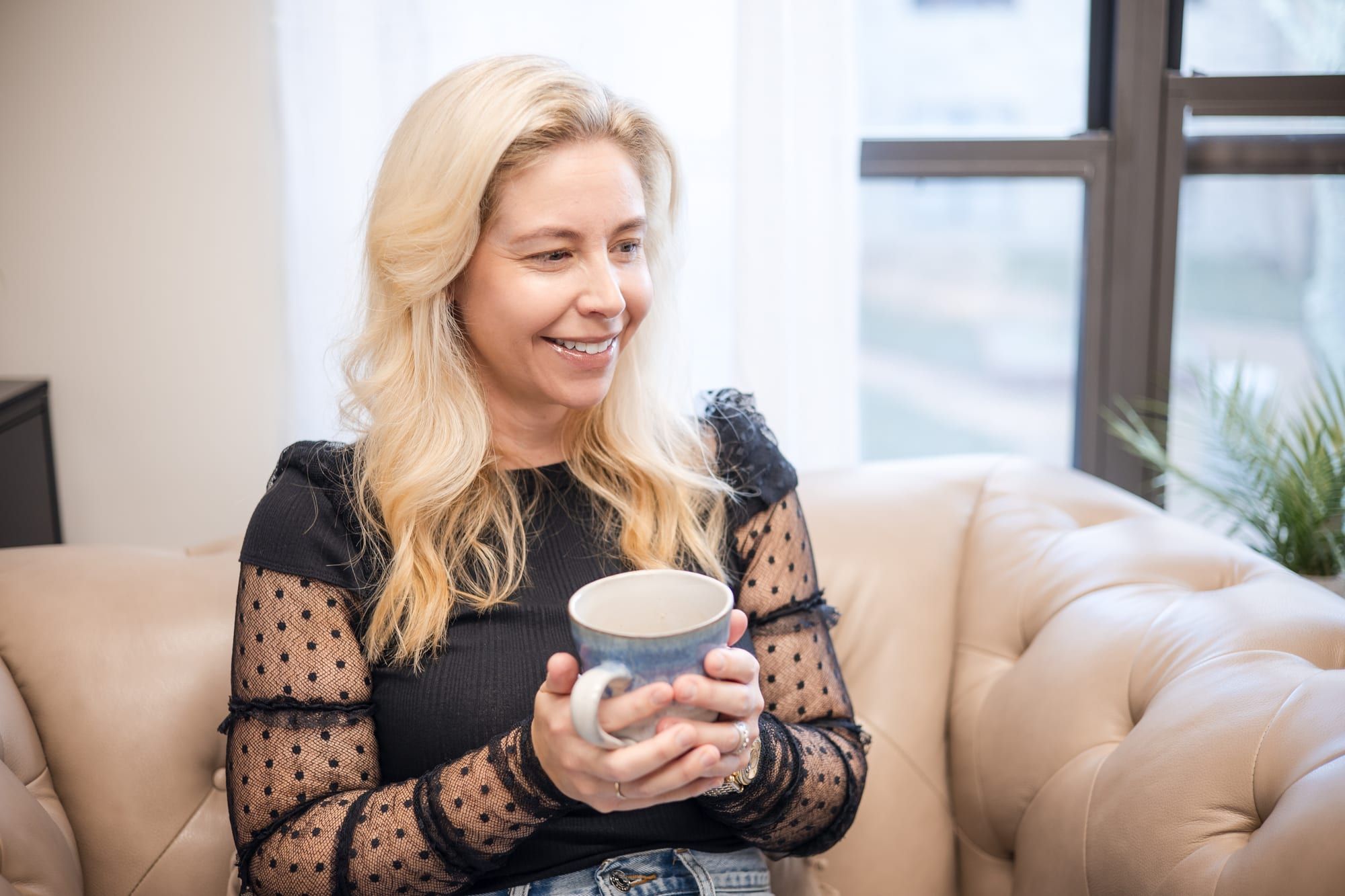 A woman with long blonde hair in a black top with sheer dotted sleeves, holding a mug and smiling as she looks toward a window from a beige leather armchair