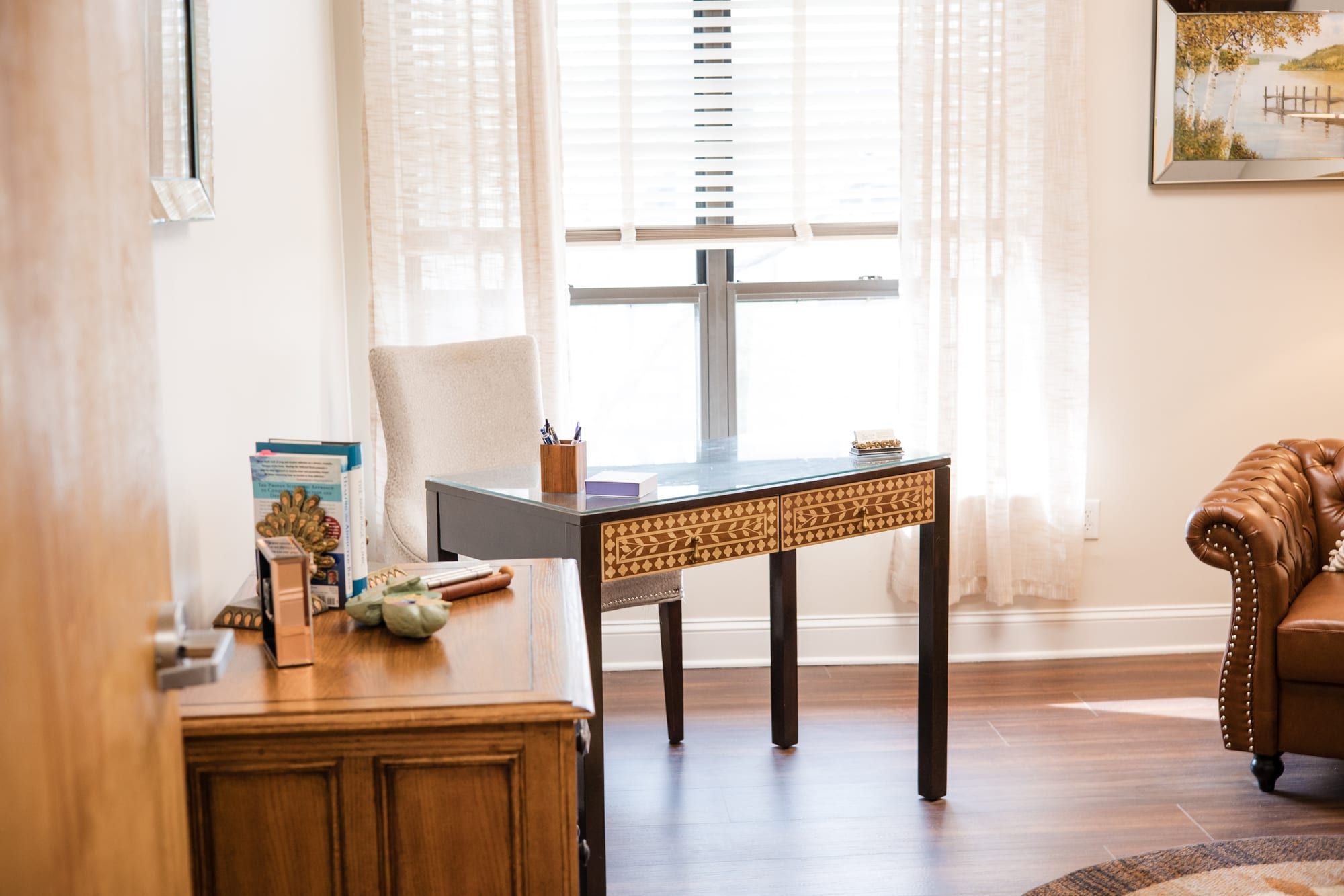 Office with a wooden desk, a brown leather armchair and a window with sheer curtains, beside a framed painting