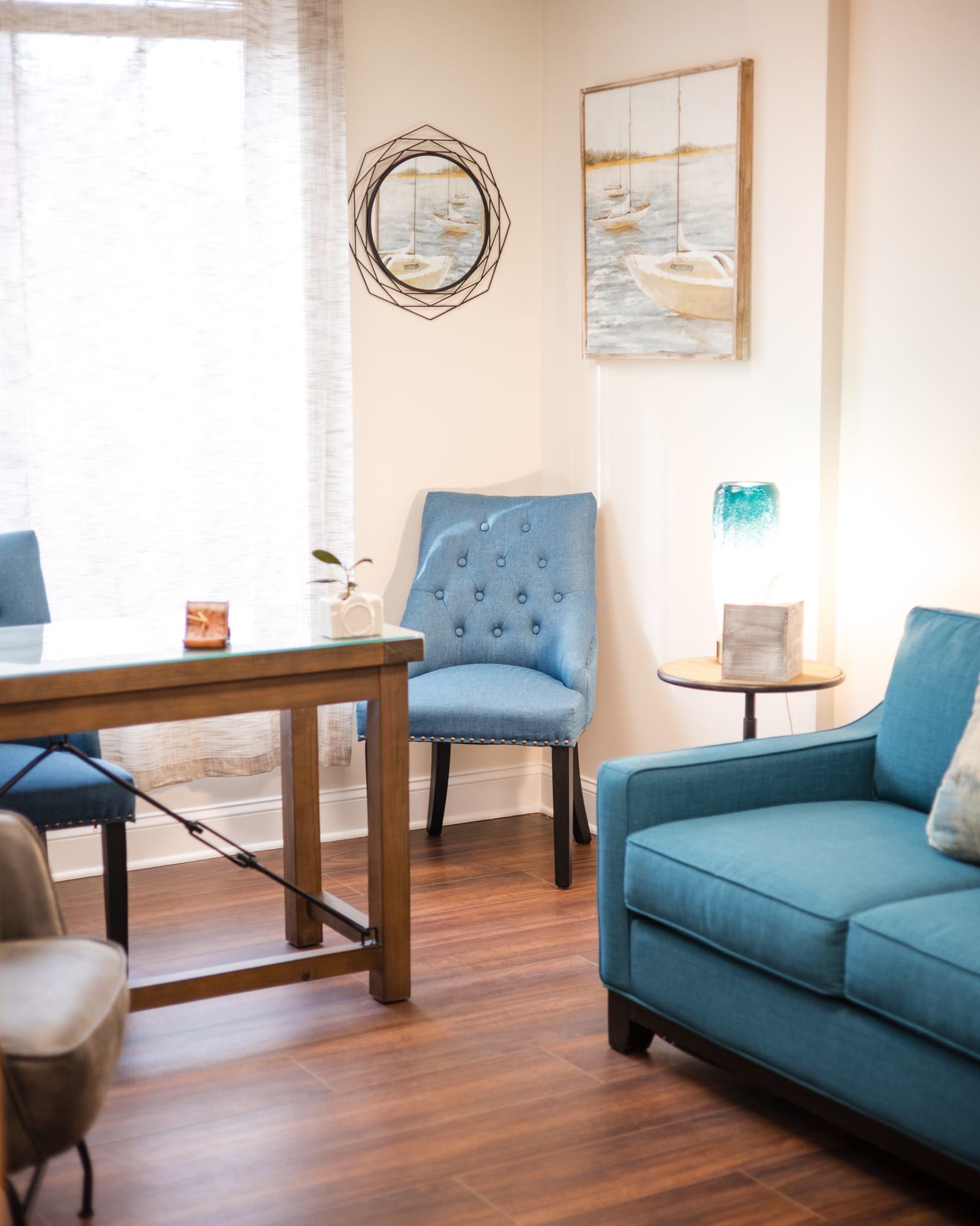 Therapy room with a teal sofa, a blue tufted chair, a wooden table and framed sailboat paintings beside a bright window