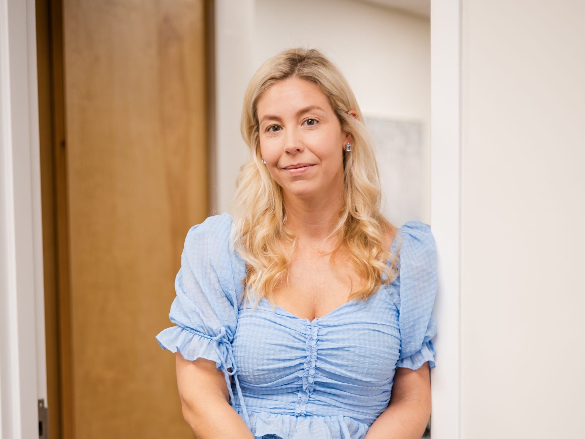 A woman with long blonde hair in a light blue ruched top, leaning in a wooden doorway with her hands clasped and a soft smile