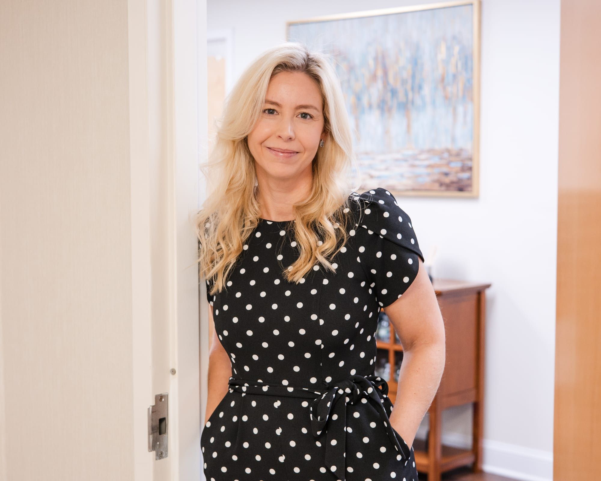A woman with long blonde hair in a black and white polka-dot jumpsuit, smiling and leaning in a wooden doorway