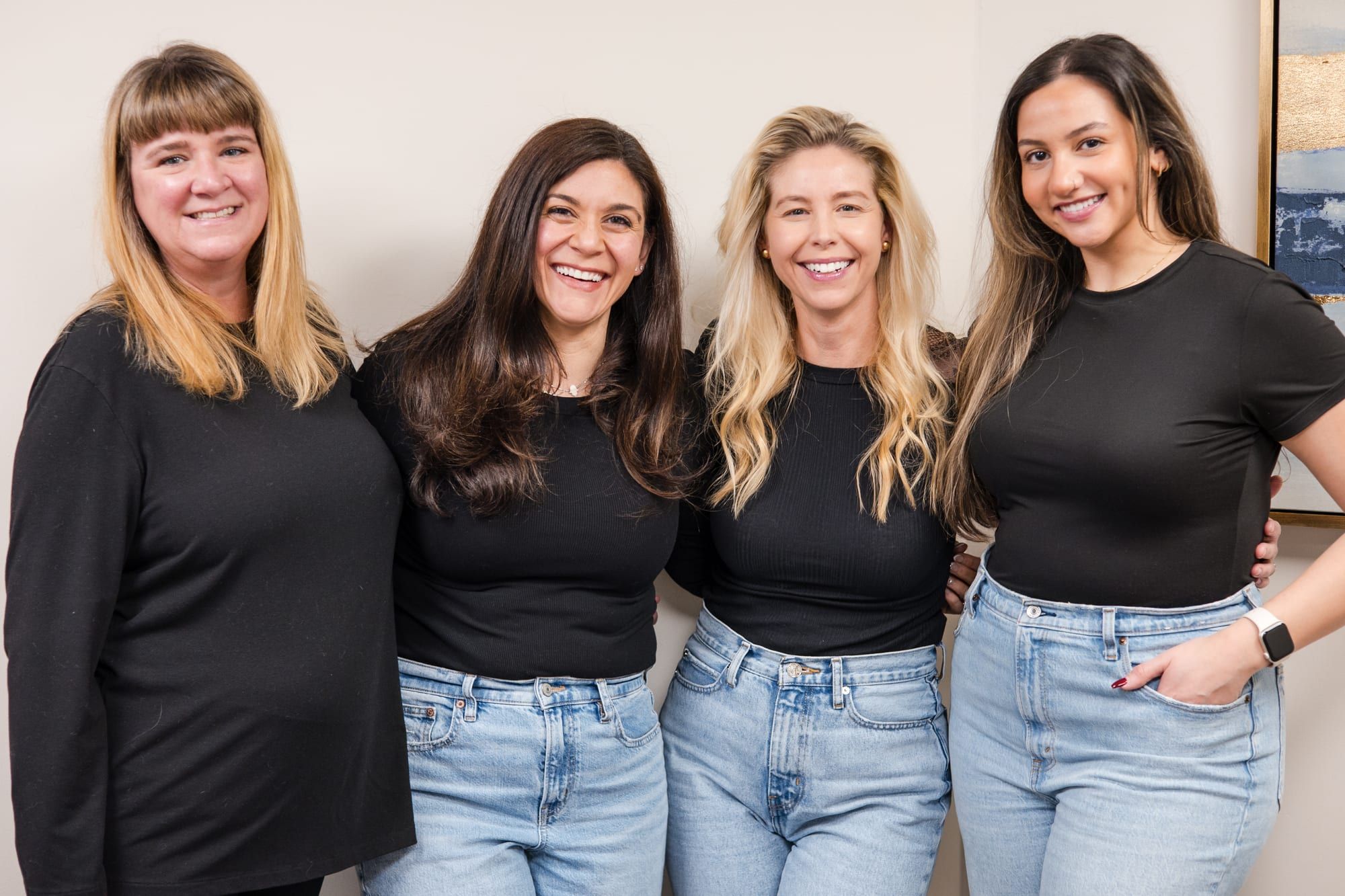Four women standing side by side with their arms around each other and smiling, in black tops and jeans