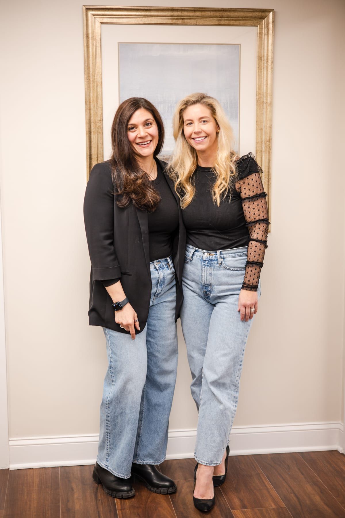 Marina Fried and Lindsey standing side by side in the office and smiling, in black tops and jeans