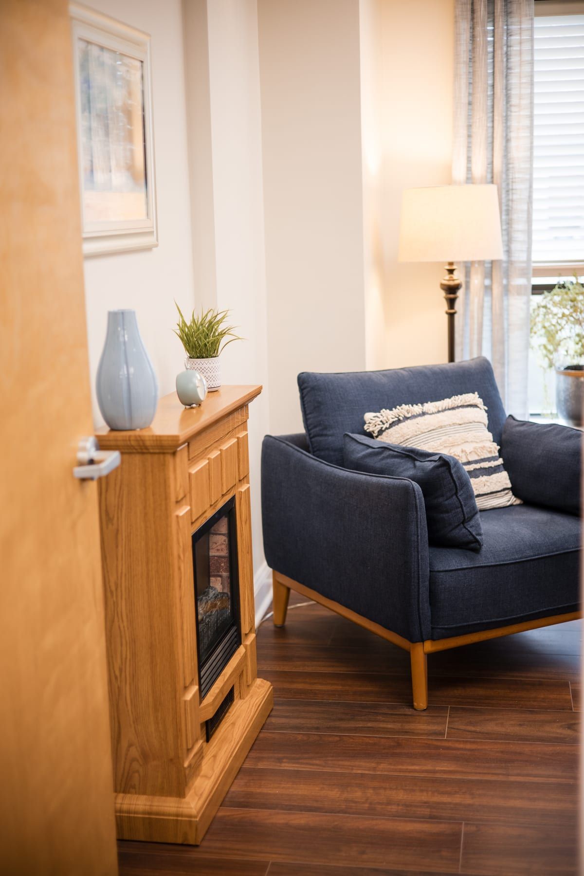 Corner of a therapy room with a navy armchair, a small wooden fireplace, a potted plant and a floor lamp by a window