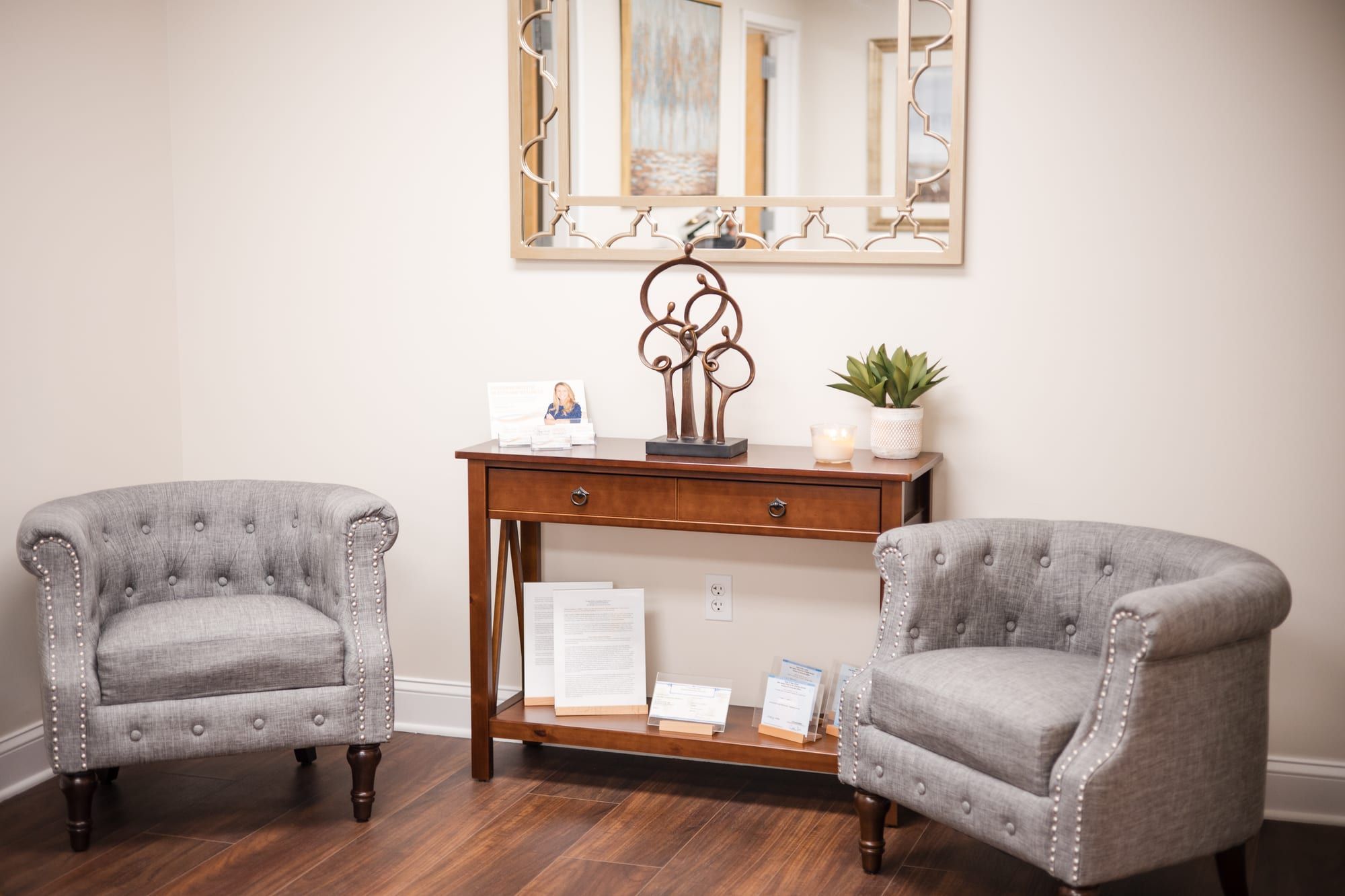 Waiting area with two grey tufted armchairs on either side of a small wooden console table, with a mirror on the wall above